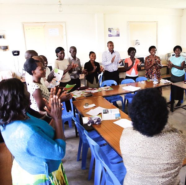 A group of adults stand in a large semi circle around a table clapping