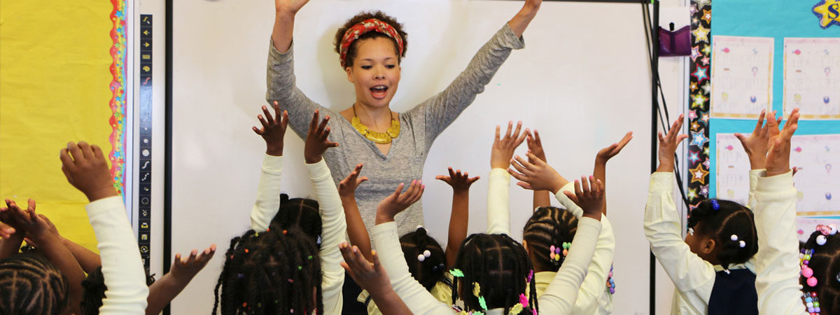 Teaching in front of classroom with hands raised, kids with hands raised