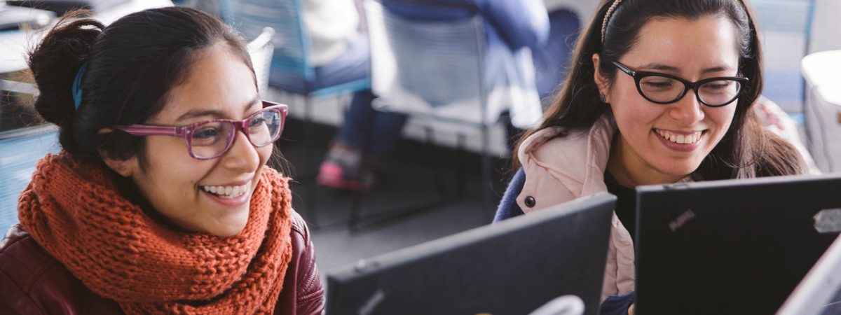 Two women sit at computers smiling and writing code