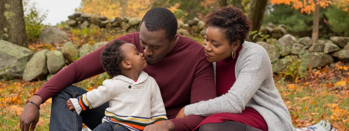 Father, mother and son sit on a picnic blanket on an autumn day. Father leans down to kiss his son lovingly.