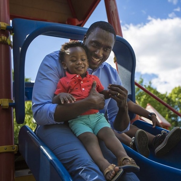 Father and son slide down a slide at a park on a sunny day.