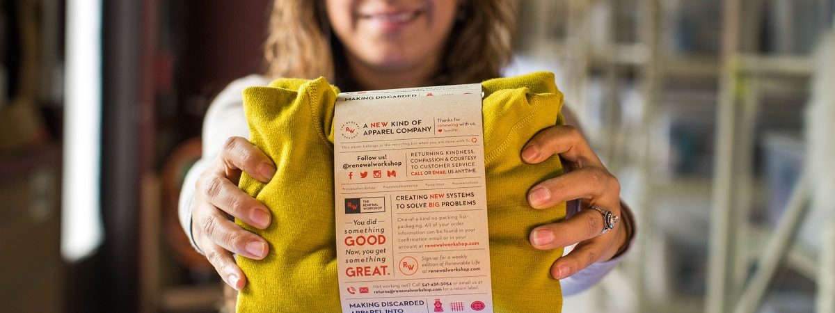 A woman holds out a yellow sweater in packaging that explains that the sweater has been repaired by The Renewal Workshop