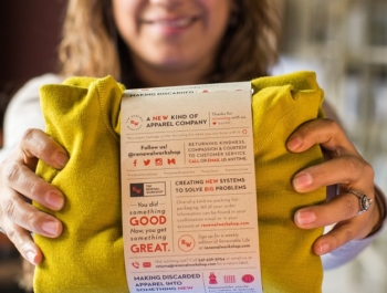 A woman holds out a yellow sweater in packaging that explains that the sweater has been repaired by The Renewal Workshop