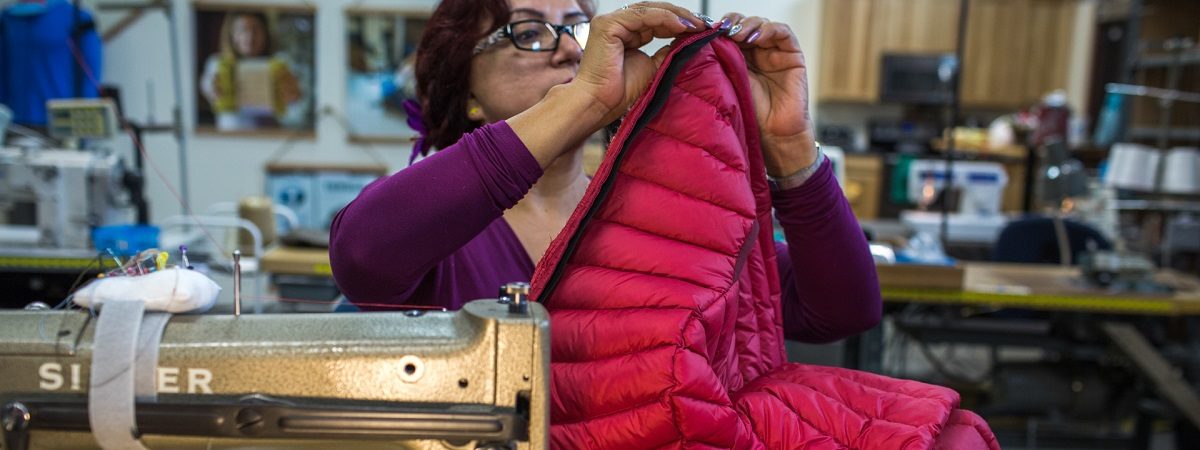 A woman sits at a sewing machine repairing a bright pink puffy jacket