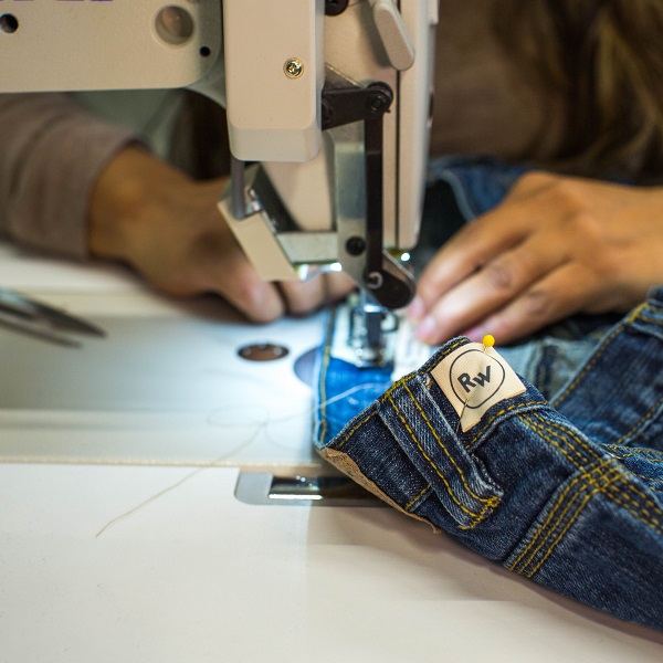 A pair of blue jeans are run through a sewing machine. The label on the jeans read