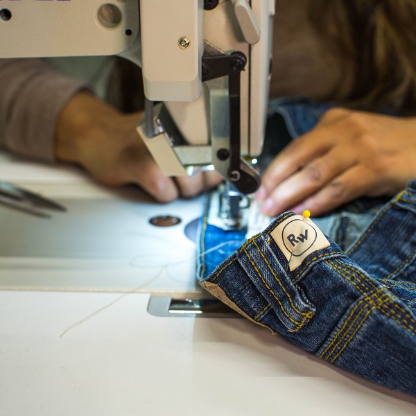 A pair of blue jeans are run through a sewing machine. The label on the jeans read 