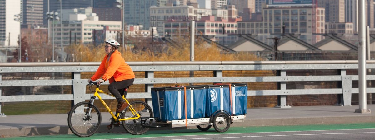 A female Wash Cycle Laundry employee riders her bike pulling a cart of laundry with the Philadelphia city scape in the background