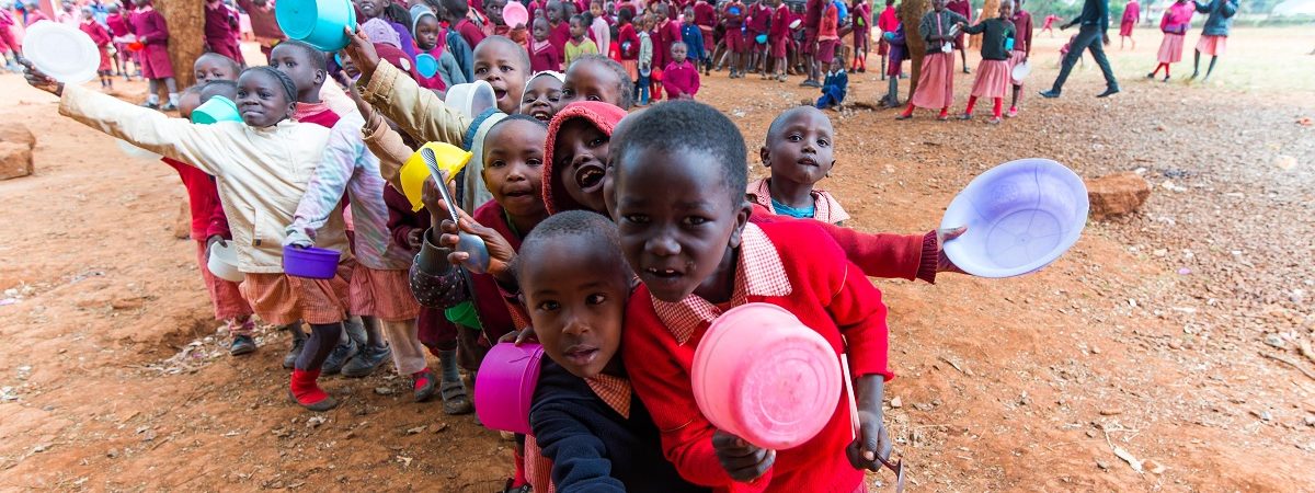 A large group of African school children wearing red uniforms gather with bowls outstretched