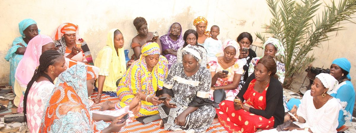 A large group of African women gather on a colorful rug and are using their mobile devices to check their savings to the MaTontine platform