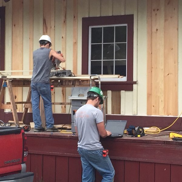 Two men in hard hats are featured; one is working to cut wood to install siding on the side of a house, while the other is using a laptop to look up parameters for the siding