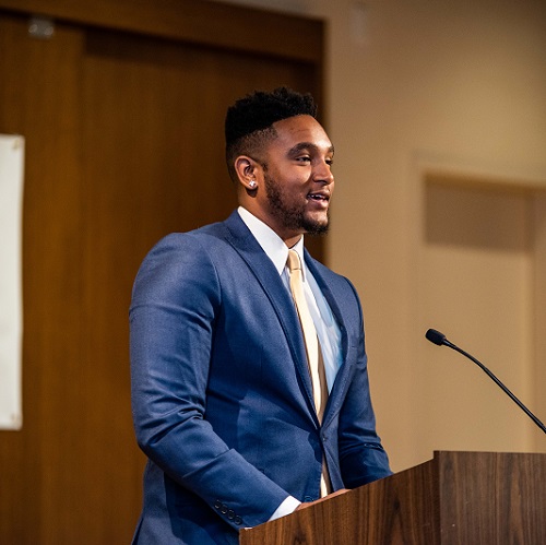 A young sharp dressed black man stands at a podium to address a crowd gathered for a Merit America event