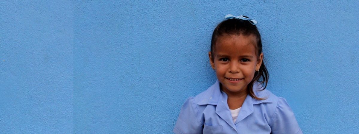 Small Costa Rican schoolgirl stands in a blue dress in front of a blue wall