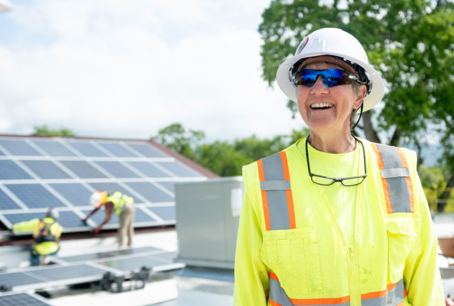 Smiling woman in a bright yellow construction vest, hard hat, and sunglasses standing in front of solar panels.