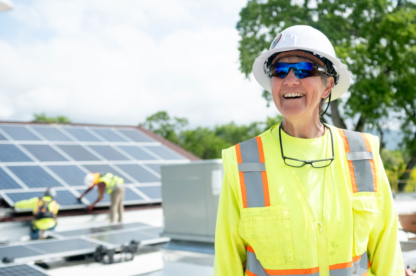 Smiling woman in a bright yellow construction vest, hard hat, and sunglasses standing in front of solar panels.