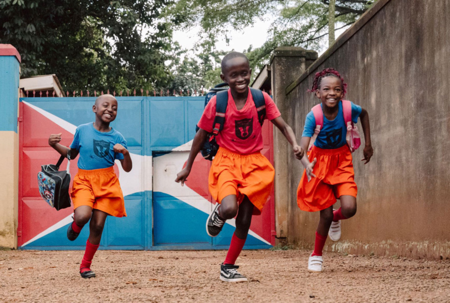 Three smiling African children wearing bright clothing and backpacks running toward camera.