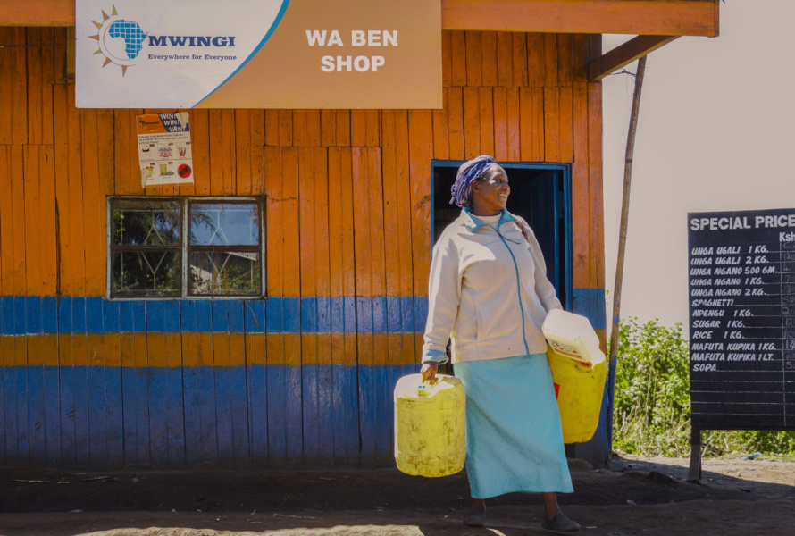 Smiling African woman holding large containers outside of a MWINGI shop.