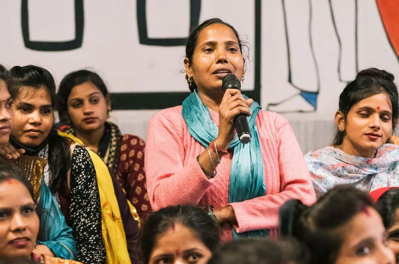 Woman dressed in a pink sweater with blue satin scarf standing amongst a seated group speaks through a microphone.