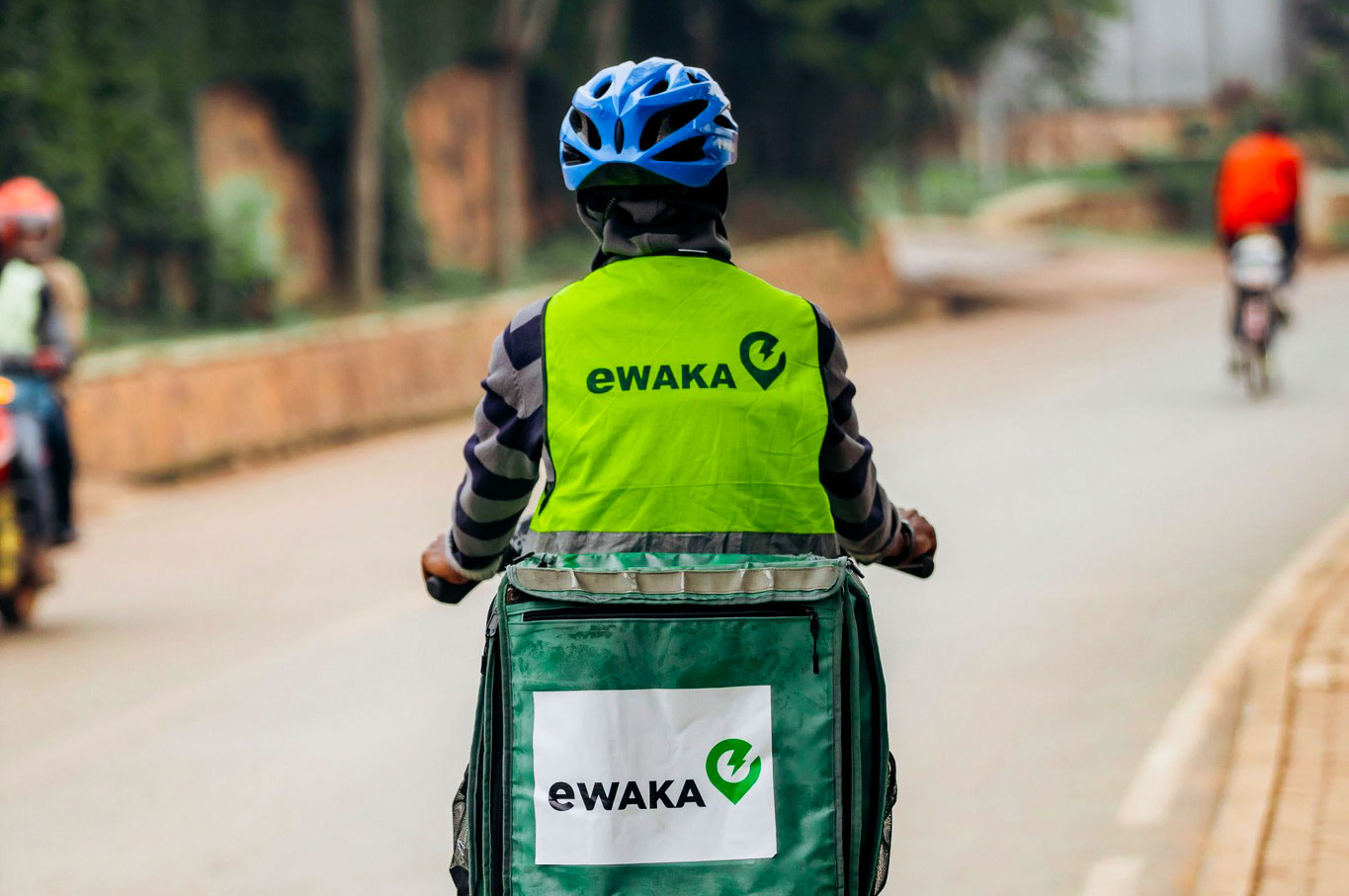 Person riding a bike wearing a bright green vest reading eWAKA across their back, and a green carrier bag reading eWAKA.