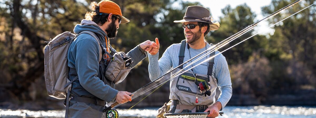 Two men stand in a river with their fishing gear and they high five each other for sharing their feelings