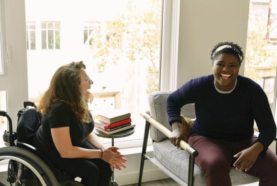 A white woman in a wheelchair laughs while sitting next to her Black friend