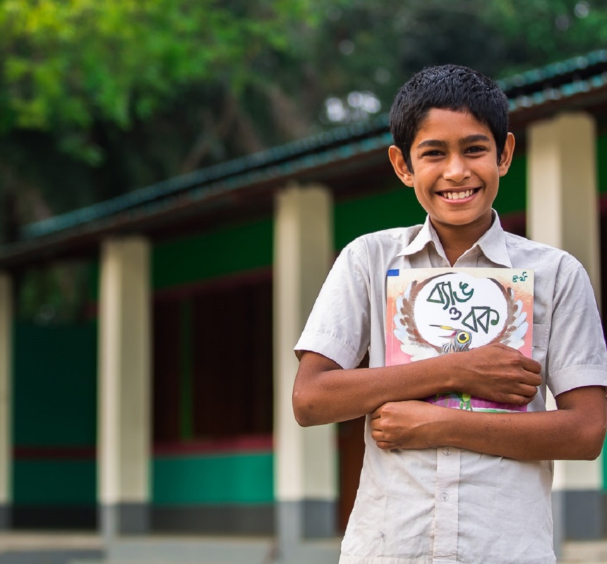 A boy smiles holding his book in front of his school