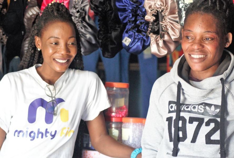Two African women smile while sitting in front of their silk hair bonnet shop.
