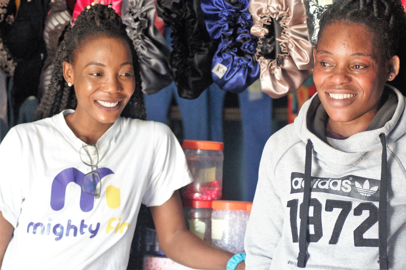 Two African women smile while sitting in front of their silk hair bonnet shop.